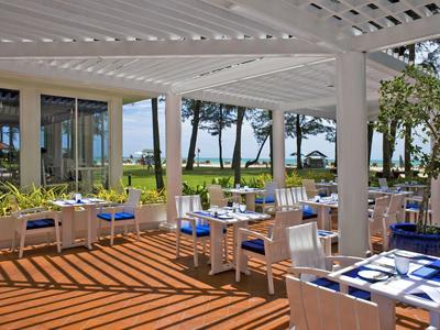 Terrasse mit weißen Stühlen und Tischen, blauer Tischdecke, überdacht mit Blick aufs Meer und Palmen.