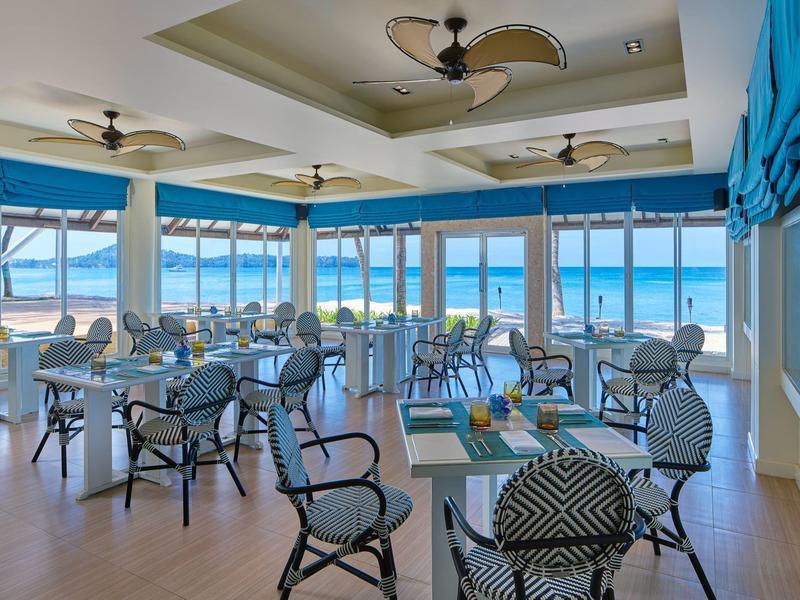 Großer, heller Speiseraum mit Glaswänden, Blick auf Meer, mehreren Stühlen und Tischen, Deckenventilatoren.