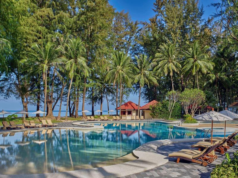 Großer Pool mit Liegestühlen, umgeben von Palmen und Bäumen am Meer an einem sonnigen Tag.