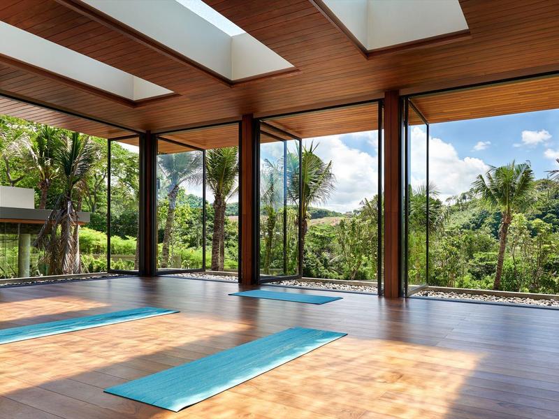 Heller Raum mit Holzboden, großen Glasfenstern und blauen Yogamatten, umgeben von Grün.