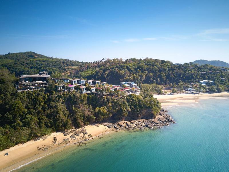 Küste mit hellem Sandstrand, klarem blauem Wasser und bewaldeten Hügeln mit Häusern darauf.