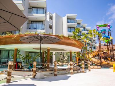 Modernes Hotel mit großem Sonnenschirm und kindgerechtem Wasserspielplatz bei sonnigem Himmel.