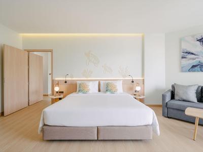 Helles Hotelzimmer mit großem Doppelbett, grauem Sofa und minimalistischer Dekoration.