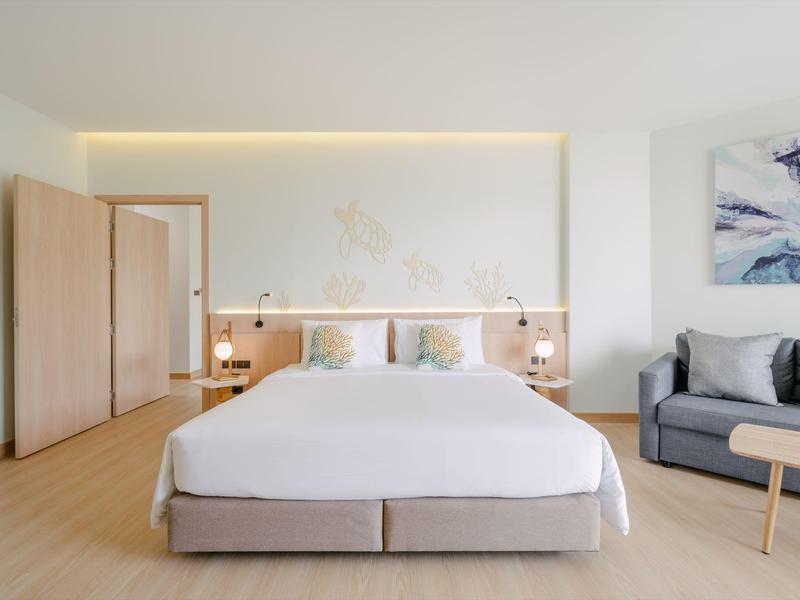 Helles Hotelzimmer mit großem Doppelbett, grauem Sofa und minimalistischer Dekoration.