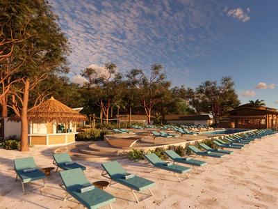 Strand mit Liegestühlen und kleinen Bungalows vor bewaldetem Hintergrund bei Sonnenuntergang.