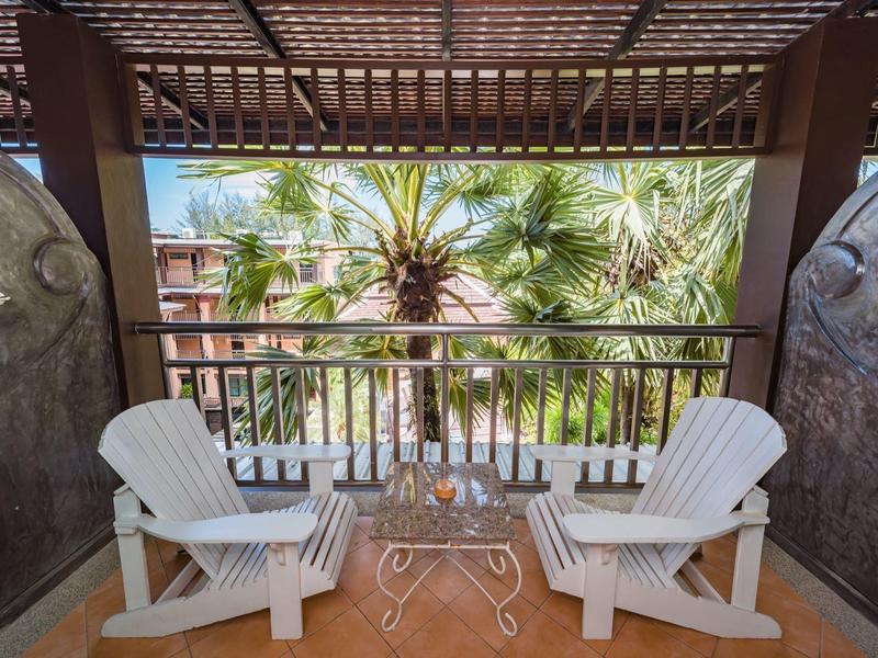 Zwei weiße Stühle mit kleinem Tisch auf einem Balkon mit Blick auf Palmen und blauen Himmel.