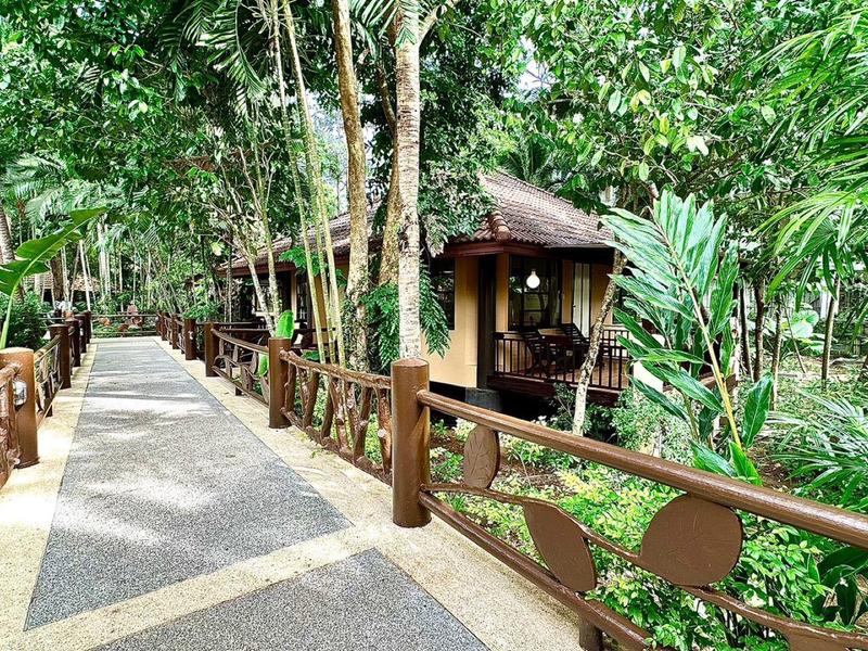 Ein walkway aus Stein mit braunen Holzgeländern führt durch einen dichten tropischen Garten zu einer kleinen Hütte.