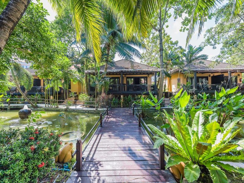 Ein Holzsteg führt durch tropischen Garten zu einem Bungalow in einem Resort.
