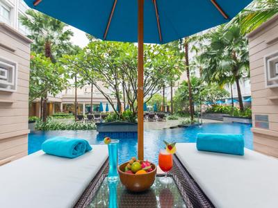 Dwa leżaki z niebieskimi poduszkami obok basenu pod niebieskim parasolem w tropikalnym otoczeniu.