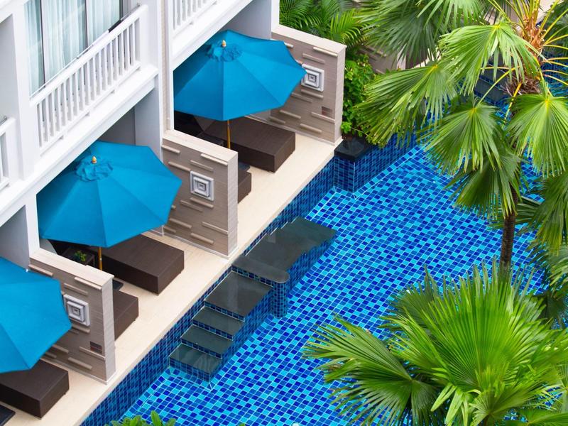 Widok na hotel z niebieskimi parasolami i leżakami obok basenu i palm.