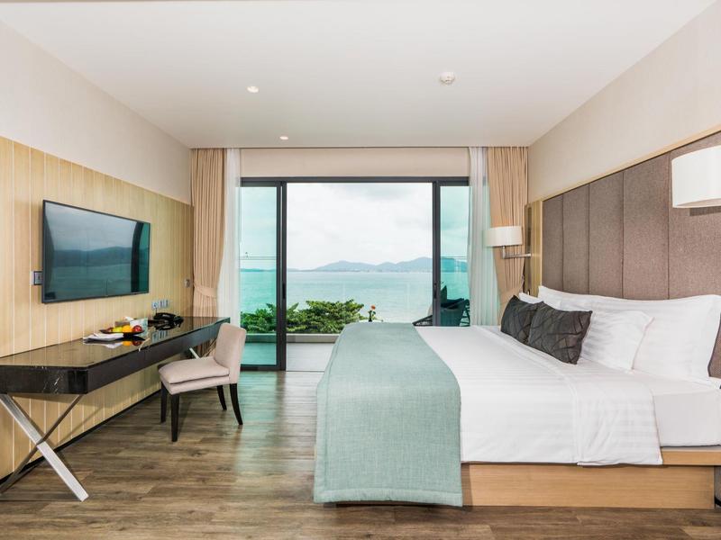 Modern hotelkamer met groot bed, bureau en zeezicht door ramen van vloer tot plafond.