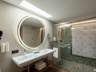 Moderní hotelová koupelna s dvojitým umyvadlem, kulatým osvětleným zrcadlem a sprchou se skleněnými dveřmi.