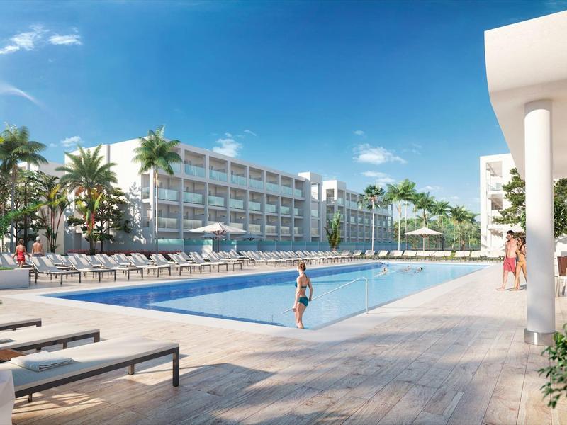 Velký venkovní bazén s lehátky, obklopený palmami a moderní hotelovou budovou pod modrou oblohou.