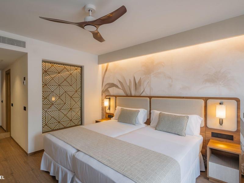 Moderní hotelový pokoj s manželskou postelí, dřevěnou podlahou a stropním ventilátorem v teplém osvětlení.