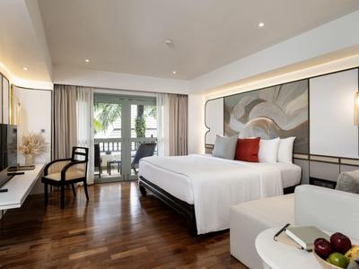 Modern eingerichtetes Hotelzimmer mit Doppelbett, Sitzecke und Balkon mit Blick auf Palmen.