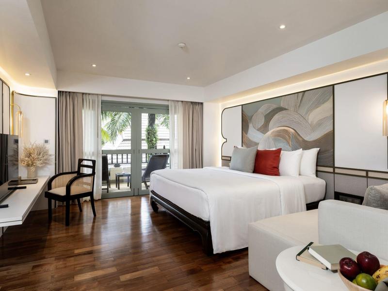 Modern eingerichtetes Hotelzimmer mit Doppelbett, Sitzecke und Balkon mit Blick auf Palmen.