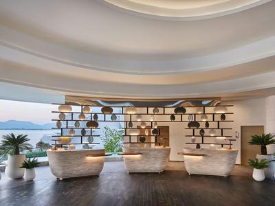 Nowoczesny salon z jasnymi fotelami, roślinami, otwartymi półkami i widokiem na morze o zachodzie słońca.