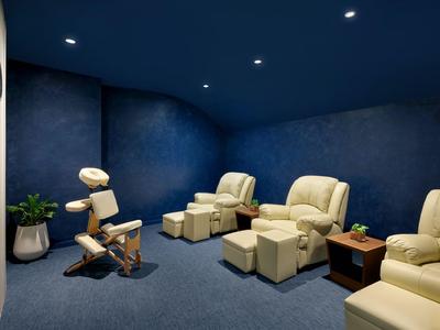 Spokojne pomieszczenie relaksacyjne z fotelem do masażu, wygodnymi fotelami i przyciemnionym niebieskim oświetleniem.