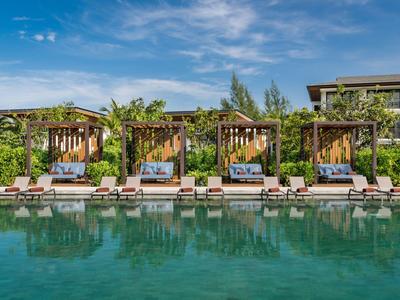 Piscine avec quatre cabanes en bois et transats devant un hôtel sous un ciel bleu.