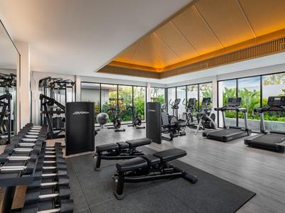 Salle de sport moderne et lumineuse avec poids, tapis de course et grandes fenêtres donnant sur la verdure.