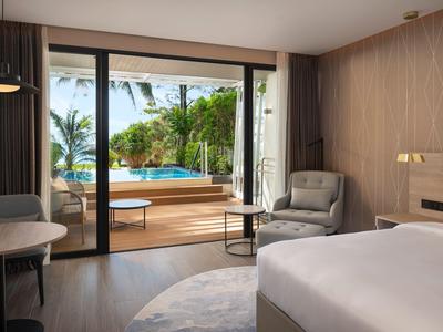 Camera d'hotel con letto matrimoniale, area salotto e accesso alla terrazza con vista su piscina e giardino.
