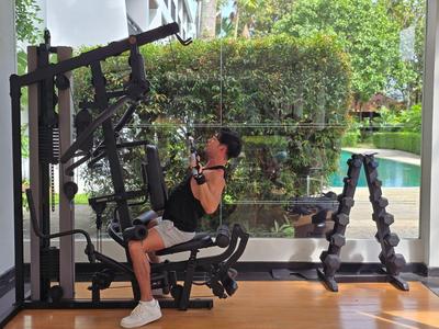 Frau trainiert mit Kabelzuggerät in hellem Fitnessraum mit Blick auf Garten und Pool.