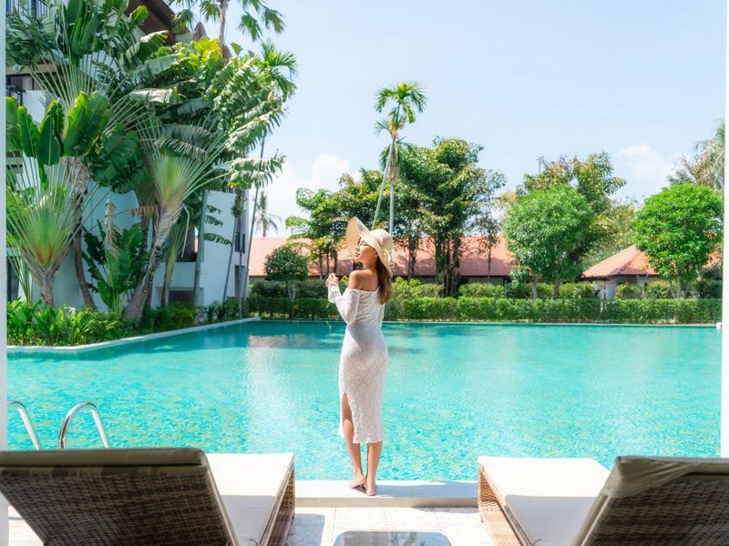 Vrouw in witte jurk staat aan de rand van een groot zwembad met tropisch groen op de achtergrond.
