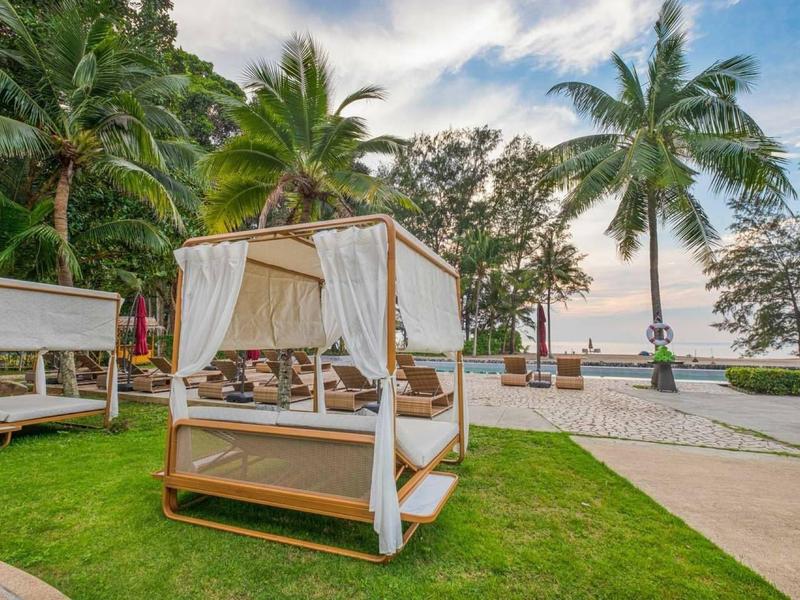 Een luxe open bed met witte gordijnen op gras voor palmbomen en zee bij zonsondergang.