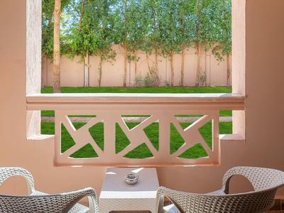 Zwei weiße Stühle und ein kleiner Tisch auf einer Terrasse mit Blick auf grüne Bäume hinter einer rosa Wand.