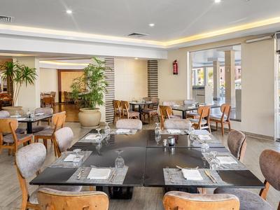 Modern restaurant with wooden tables and chairs set for guests, bright lighting, and plants in the background.