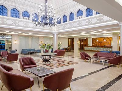 Bright hotel lobby with elegant seating areas and a chandelier under a glass roof.