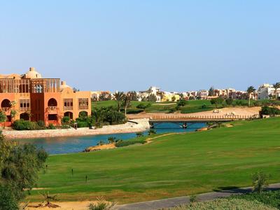 Grüner Golfplatz mit Teich, Brücke und orangefarbenem Gebäude unter blauem Himmel.