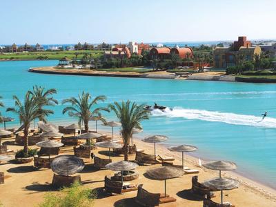 Strand mit Palmen, Strohhütten und blauem Wasser, im Hintergrund Gebäude und klare Himmel.