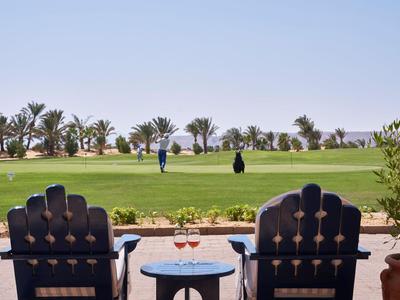 Two wooden chairs and a table with two wine glasses in front of a golf course with players in the background.