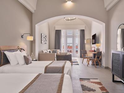 Helles Hotelzimmer mit zwei Doppelbetten, Sofa, Schreibtisch, Balkon und modernem Interieur.