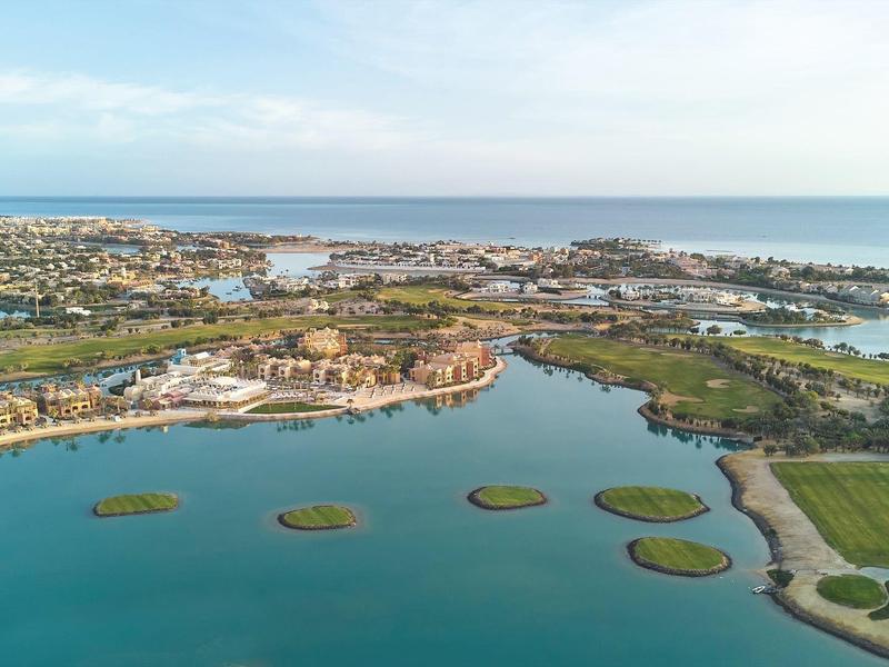 Luftaufnahme einer Küstenlandschaft mit blauem Wasser, Inseln, grünen Feldern und Gebäuden im Hintergrund.