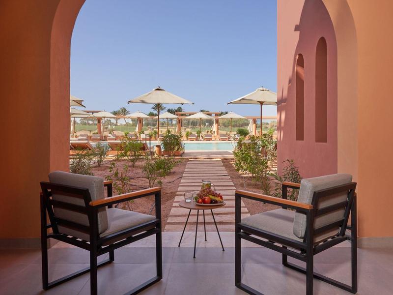 Zwei moderne Stühle mit Kissen vor Bögen, Blick auf Terrasse mit Pflanzen und Sonnenschirmen.