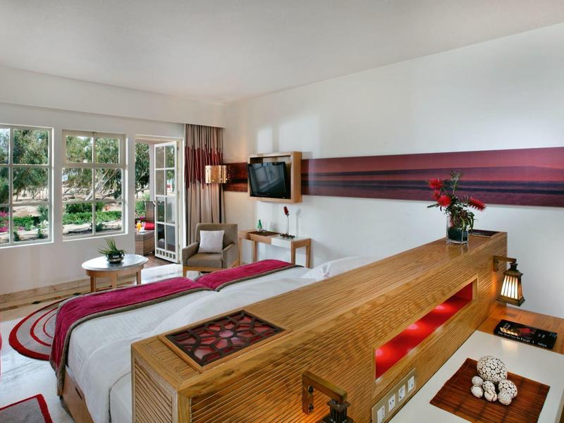 Modern hotel room with a large bed, red and white decor, and a window overlooking a pool area.