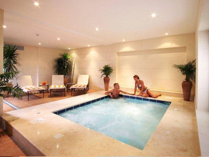 Innenraum mit kleinem quadratischem Pool, zwei Personen am Beckenrand, warme Beleuchtung, Pflanzen.