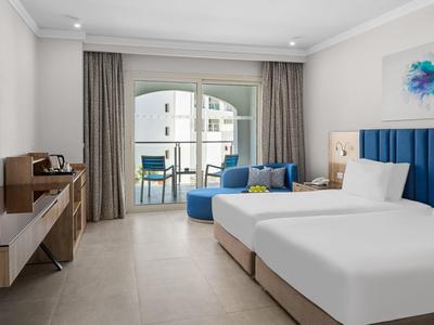 Modernes Hotelzimmer mit Doppelbett, blauem Sofa, TV, Balkon und hellem Parkettboden.