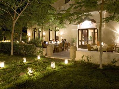 Garten mit Bäumen und Laternen, Gäste sitzen abends auf Terrasse eines beleuchteten Hauses.