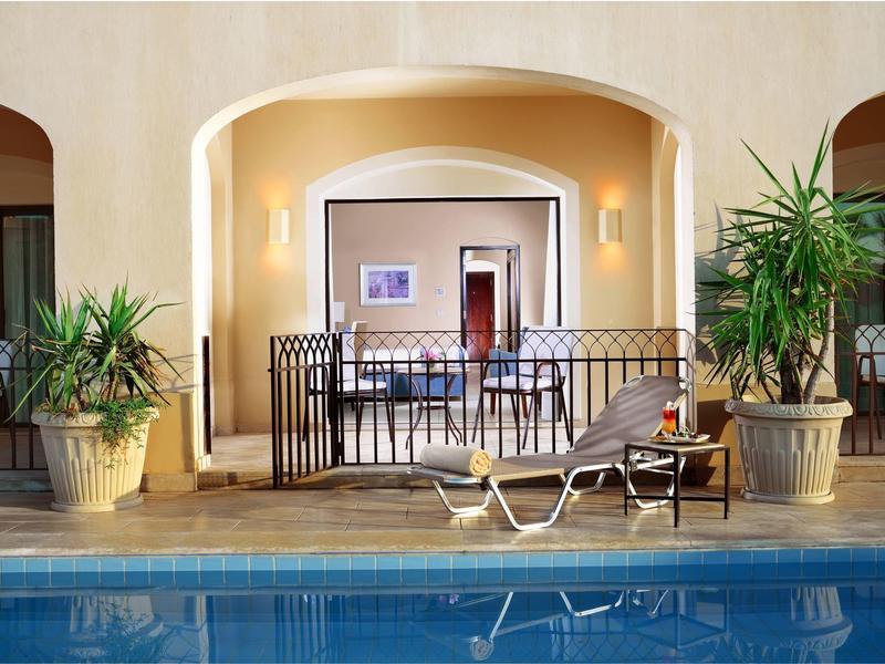 Schwimmbecken mit Liegestuhl, Tischen, großen Pflanzen und geschwungenen Bögen im Hintergrund.