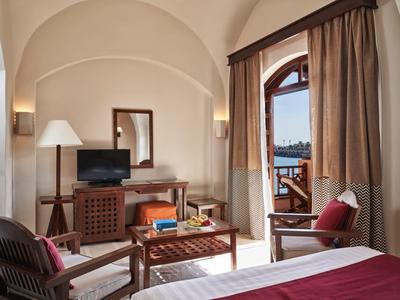 Helles Hotelzimmer mit hohen gewölbten Decken, Holzmöbeln und Blick auf Meer durch Balkon.