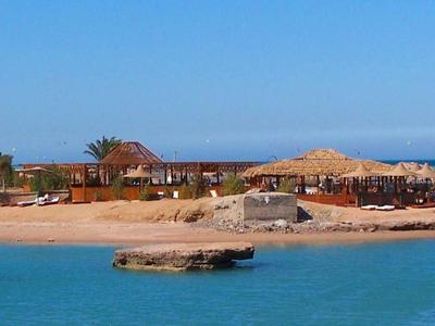 Strand mit klarem blauem Wasser, Sand, Palmen und Gebäuden unter blauem Himmel.