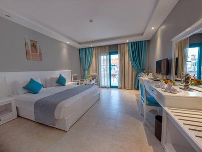 Helles Hotelzimmer mit Doppelbett, blauen Kissen, Schreibtisch, Fernseher und Balkon mit Vorhängen.