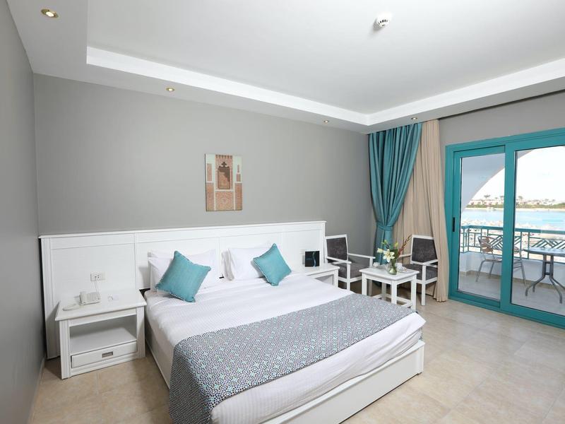 Helles Zimmer mit großem Bett, grauen Wänden, türkisfarbenen Vorhängen und Balkon mit Meerblick.