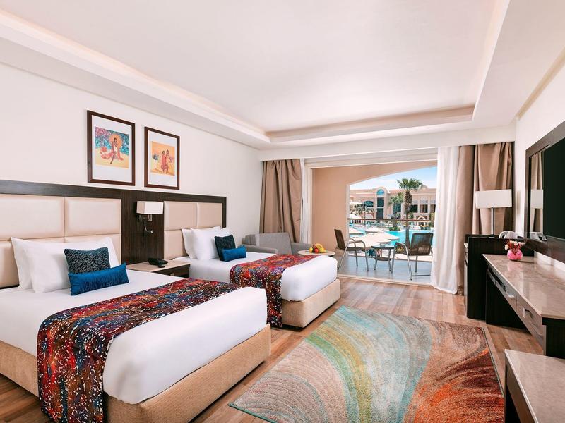 Hotel room with two beds facing a TV and a balcony overlooking a pool area.