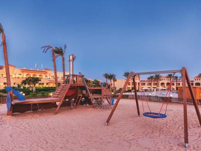 A sandy children's playground with swing and climbing frame in front of a hotel under a blue sky.