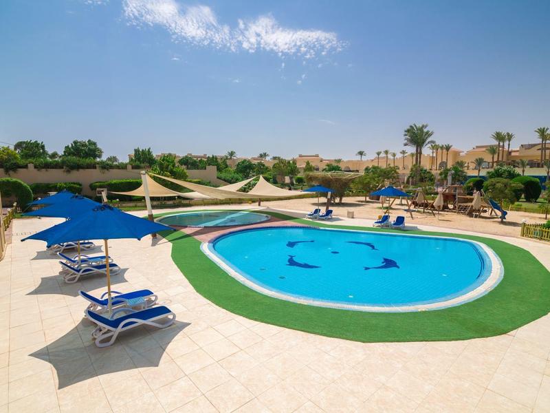 Round pool with blue loungers and umbrellas in a sunny hotel area.