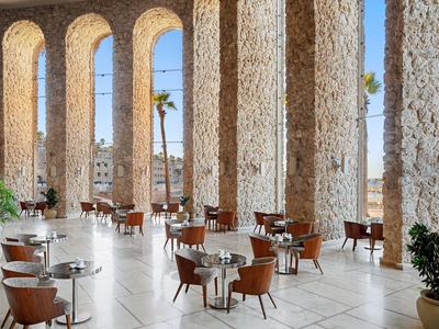 Area pranzo aperta con tavoli rotondi e sedie tra alte colonne alla luce del giorno.